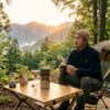 「日本人の男性がキャンプ場の朝日の中で、ポータブルコーヒーメーカーで淹れたコーヒーを楽しみながら静かに過ごしている様子。キャンプや車中泊でのコーヒー時間の格別さを表現した、自然に囲まれた朝の風景」