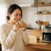 朝の光の中でマグカップのコーヒーを両手で包み、目を閉じて味わう日本人女性。