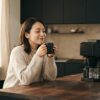 マットブラックの全自動コーヒーメーカーとガラスカラフェが置かれた明るいモダンミニマリストのキッチンで、温かいセラミックマグを両手で包み込みながら満足そうに微笑む30代の日本人女性の朝のコーヒータイム風景