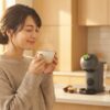 朝陽が差し込む明るいキッチンで、温かいコーヒーを両手で穏やかに抱える日本人女性の後ろにドルチェグスト本体が見える、ドルチェグストのある朝の始まりを表現するアイキャッチ画像。