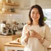 明るいミニマルなキッチンで淹れたてのコーヒーを笑顔で楽しむ30代の日本人女性。朝陽が差し込む背景に、コーヒーメーカーが柔らかくぼかされている温かなライフスタイル写真。