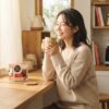 朝の柔らかな光が差し込むキッチンで、温かいマグカップを両手で包む日本人女性。カウンターには色鮮やかなデザインの、コンパクトなドルチェグストのカプセルボックスが置かれている朝のシーン。