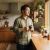 朝の木製カウンターで、磁器カップを手に窓から差し込む光を見つめる日本人男性のリラックスした朝のひととき