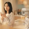 朝の光が差し込む明るいキッチンでマグカップを手にリラックスした笑顔の日本人女性。背景にキューリグのコーヒーメーカーがわずかに見えるライフスタイル写真。