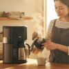 朝の日差しが入るキッチンで、湯気の立つコーヒーメーカーを眺めながら挽きたてのコーヒーを味わう日本人女性の笑顔